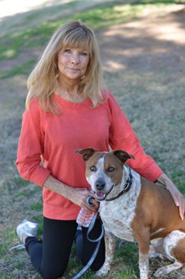 A woman in a red top and black pants kneeling next to a smiling brown and white dog in a park.